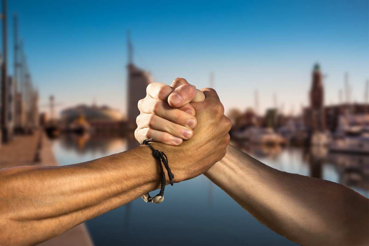 Abgebildet sind zwei Hände in einem kräftigen Handschlag zur Begrüßung. Im Hintergrund ist unscharf die Stadt Bremerhaven zu erkennen.