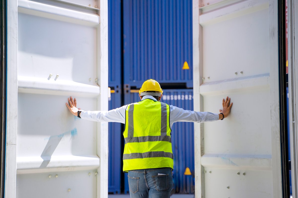 Das Bild zeigt einen Mann in greller Warnweste und Helm, der einen großen Schiffscontainer von innen her öffnet.